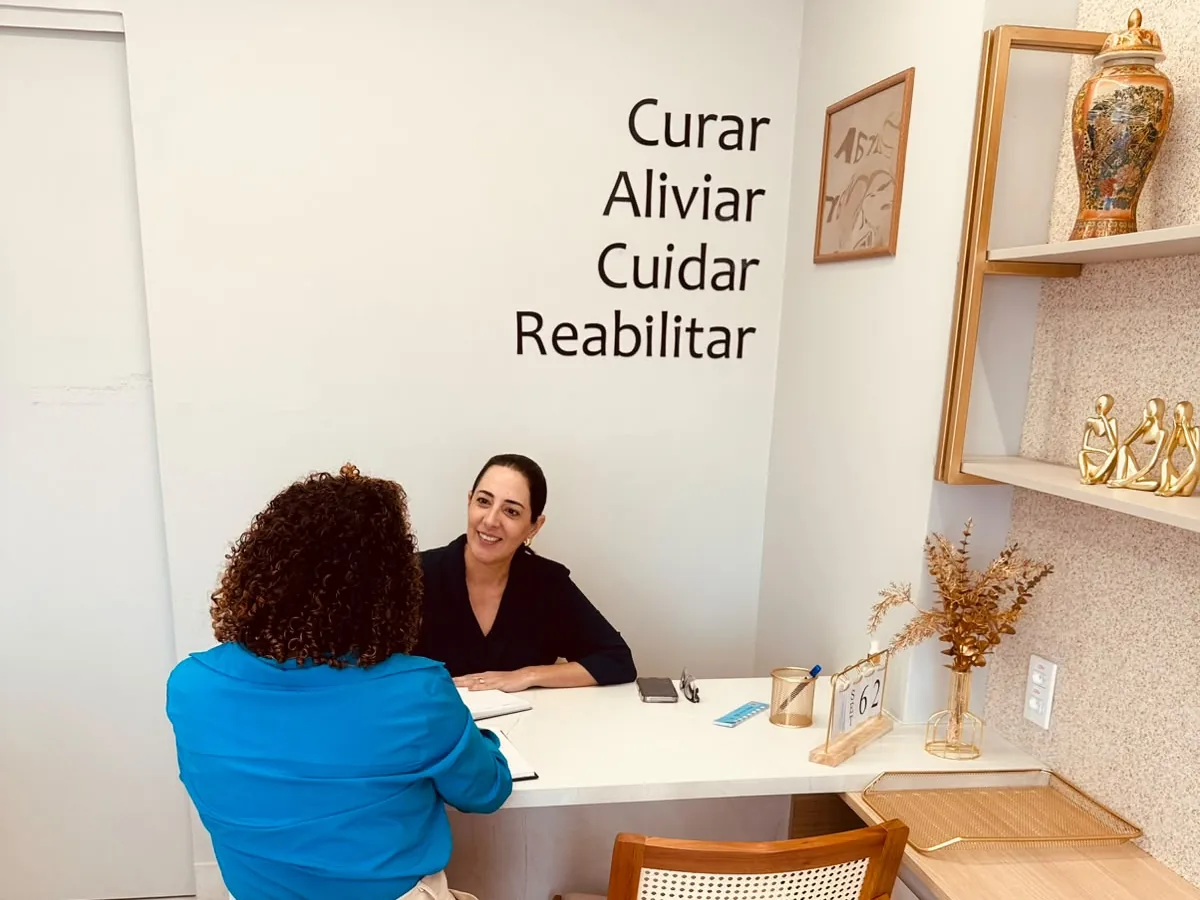 Taisa em consulta na sala de atendimento, com a frase Curar, Aliviar, Cuidar, Reabilitar na parede